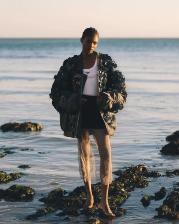 Model standing on rocky shoreline wearing embellished bomber jacket and sheer skirt by photographer Morgan Roberts