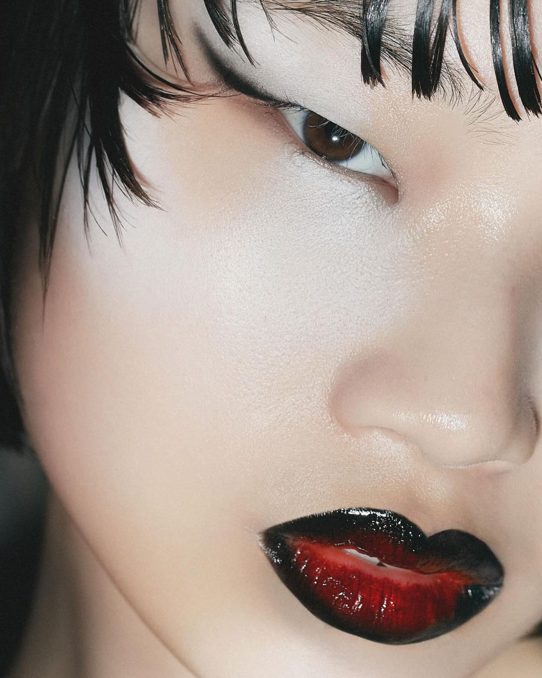Close-up of a young fair-skinned woman's face featuring dark graphic eye makeup and red to black ombre lip color and wet-look hair.