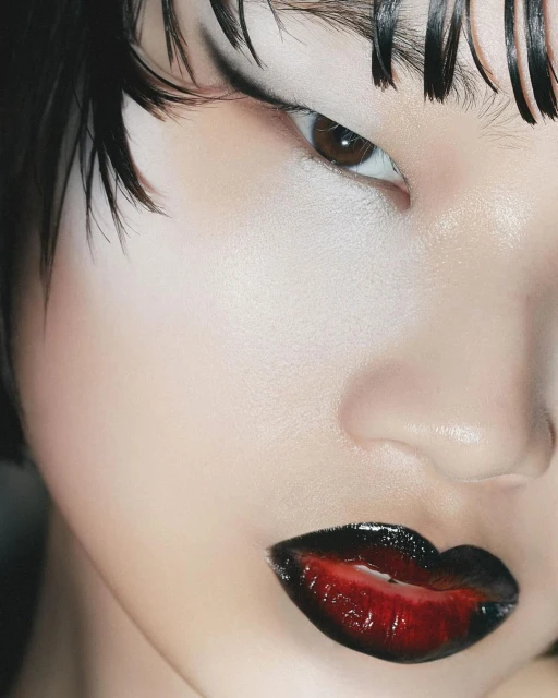 Close-up of a young fair-skinned woman's face featuring dark graphic eye makeup and red to black ombre lip color and wet-look hair.