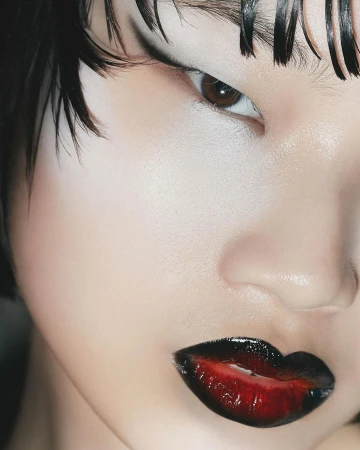 Close-up of a young fair-skinned woman's face featuring dark graphic eye makeup and red to black ombre lip color and wet-look hair.
