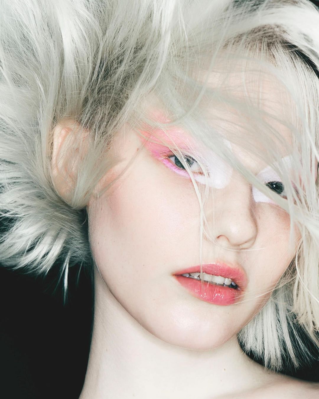 Close-up portrait of a young woman with fair skin, striking pink eye makeup, and voluminous, silver hair against a dark background.