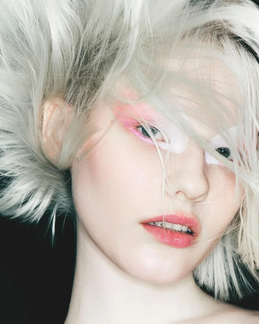 Close-up portrait of a young woman with fair skin, striking pink eye makeup, and voluminous, silver hair against a dark background.