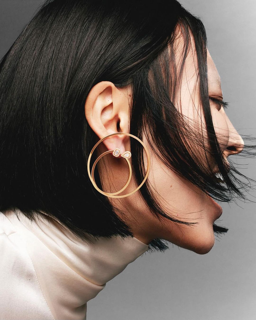 Profile portrait of an adult woman with dark hair wearing large golden hoop earrings and a light-colored turtleneck sweater.