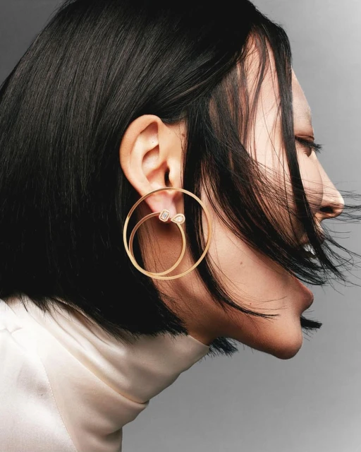 Profile portrait of an adult woman with dark hair wearing large golden hoop earrings and a light-colored turtleneck sweater.