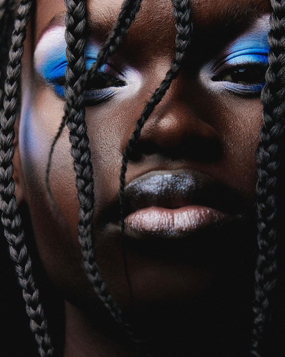 Close-up portrait of a dark-skinned woman with vibrant blue and white eye makeup and braided hair framing her face, on a dark backdrop.