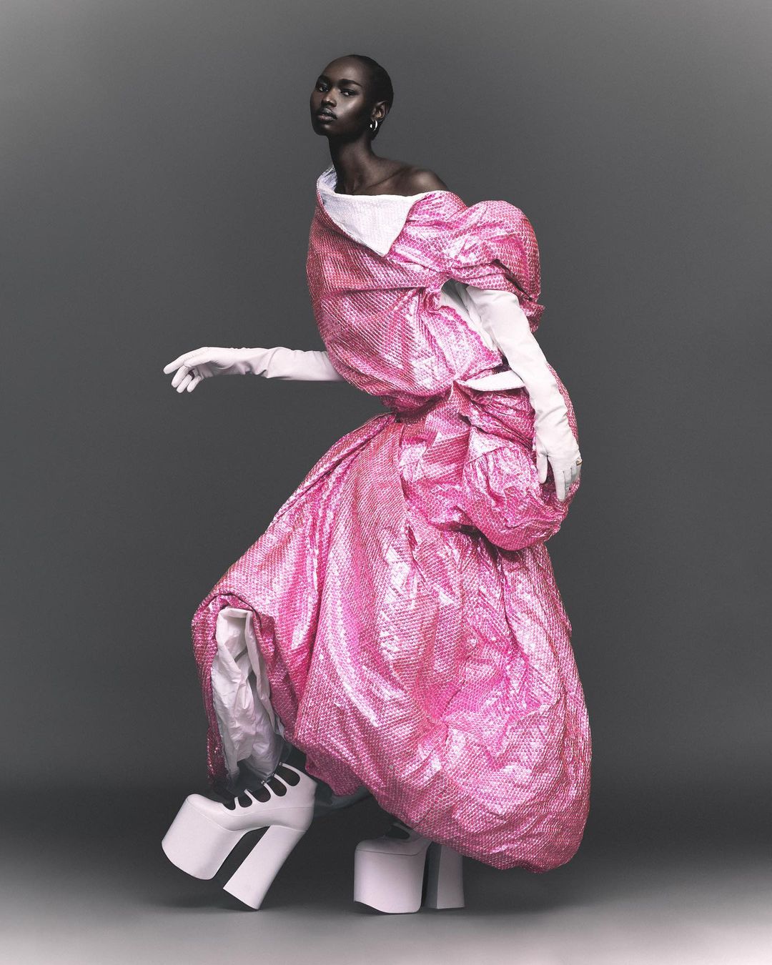 Fashion editorial image: A slender black woman in a pink dress and white platform heels poses against a gray backdrop in a photo studio.