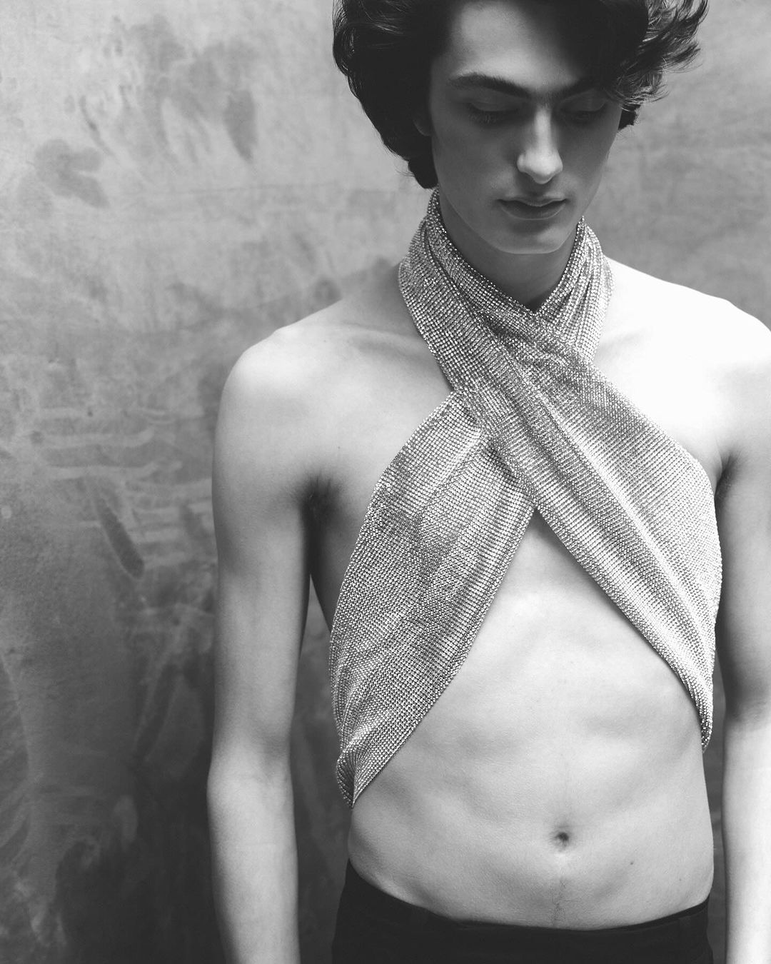 Monochrome portrait of a shirtless male model with dark hair wearing a chainmail top, posing against a textured wall.