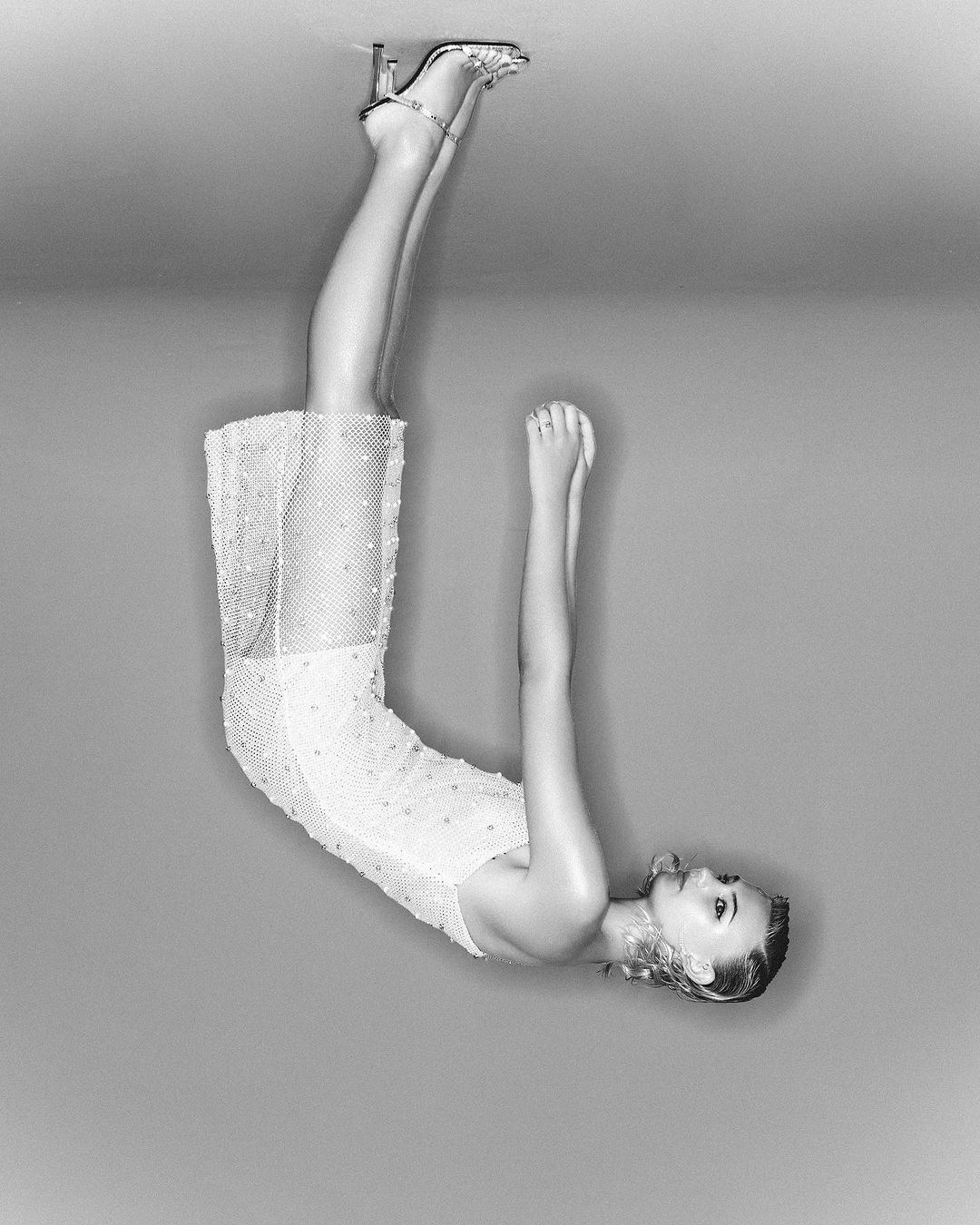 Monochrome portrait of a woman in a white dress and silver high heels, positioned upside down against a neutral gray backdrop. 