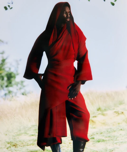 A dark-skinned fashion model poses in an earthy outdoor setting, wearing a striking red hooded outfit with flowing sleeves and wide-legged pants.