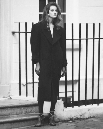 A woman in a long black coat and strapped heels standing outdoors by metal railings in front of a building.