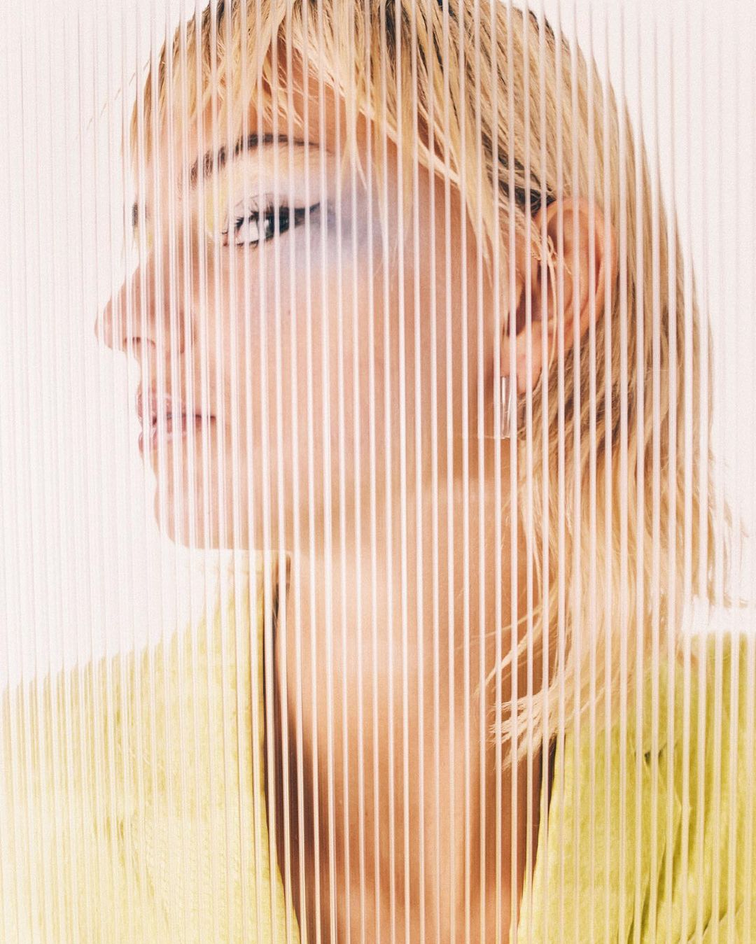 Close-up side portrait of a blonde adult female wearing a yellow blazer, partially obscured by vertical transparent lines against a white background.