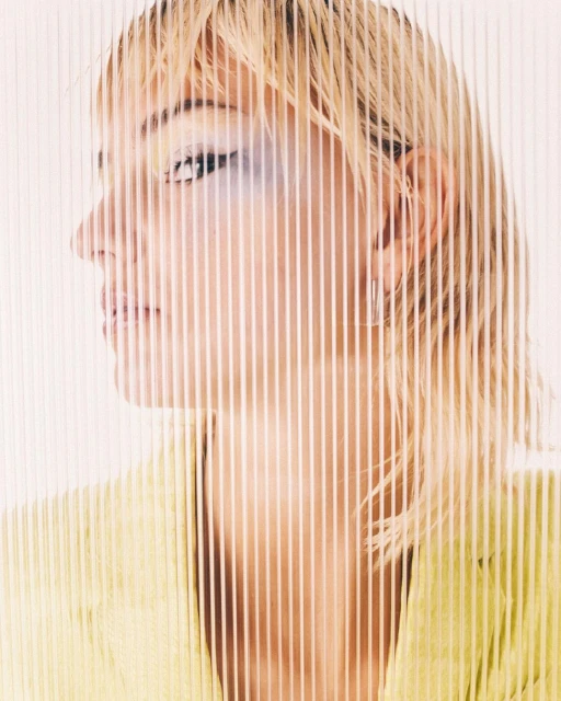 Close-up side portrait of a blonde adult female wearing a yellow blazer, partially obscured by vertical transparent lines against a white background.
