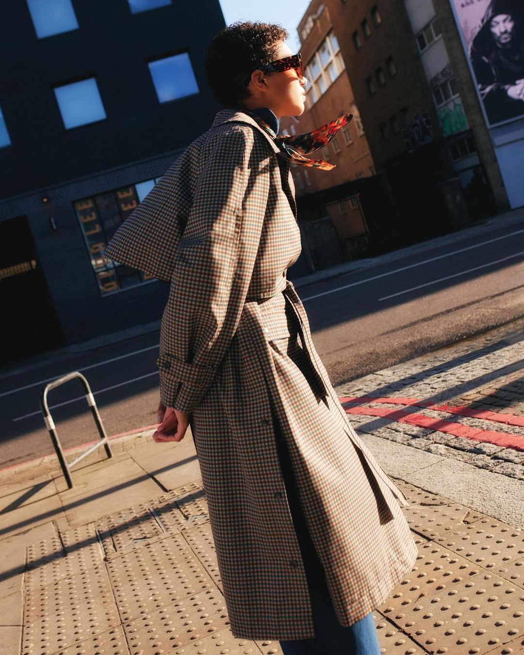 Model in houndstooth trench coat and sunglasses walking down a London street in sunlight