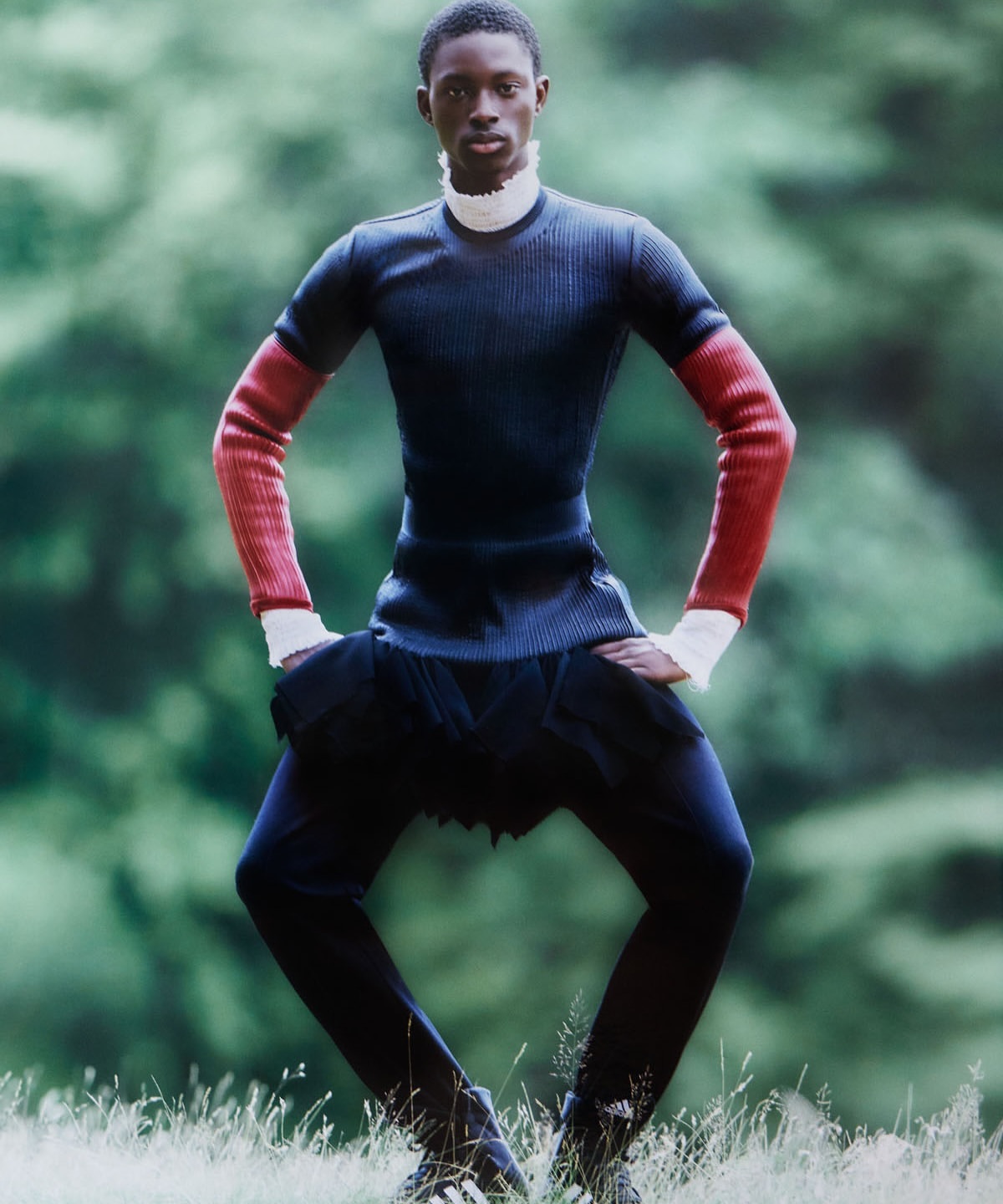 A slender black man in a ribbed, color-blocked top and dark trousers poses in a grassy field with a blurred green forest backdrop.