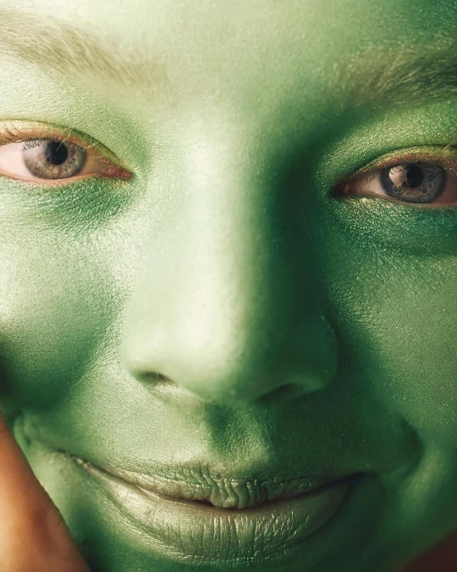 Close-up of a woman's face covered in green body paint, highlighting her eyes, nose, and lips with an overall vibrant, monochromatic effect.