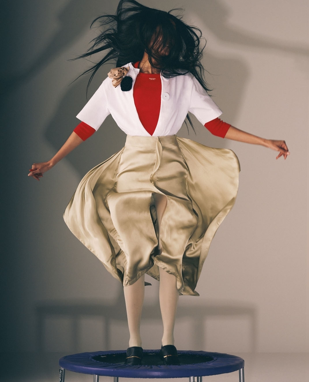 A woman in mid-air on a trampoline, wearing a red top, white jacket, and light yellow satin skirt, with her hair flying around her head.