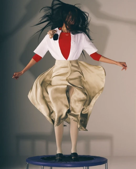 A woman in mid-air on a trampoline, wearing a red top, white jacket, and light yellow satin skirt, with her hair flying around her head.