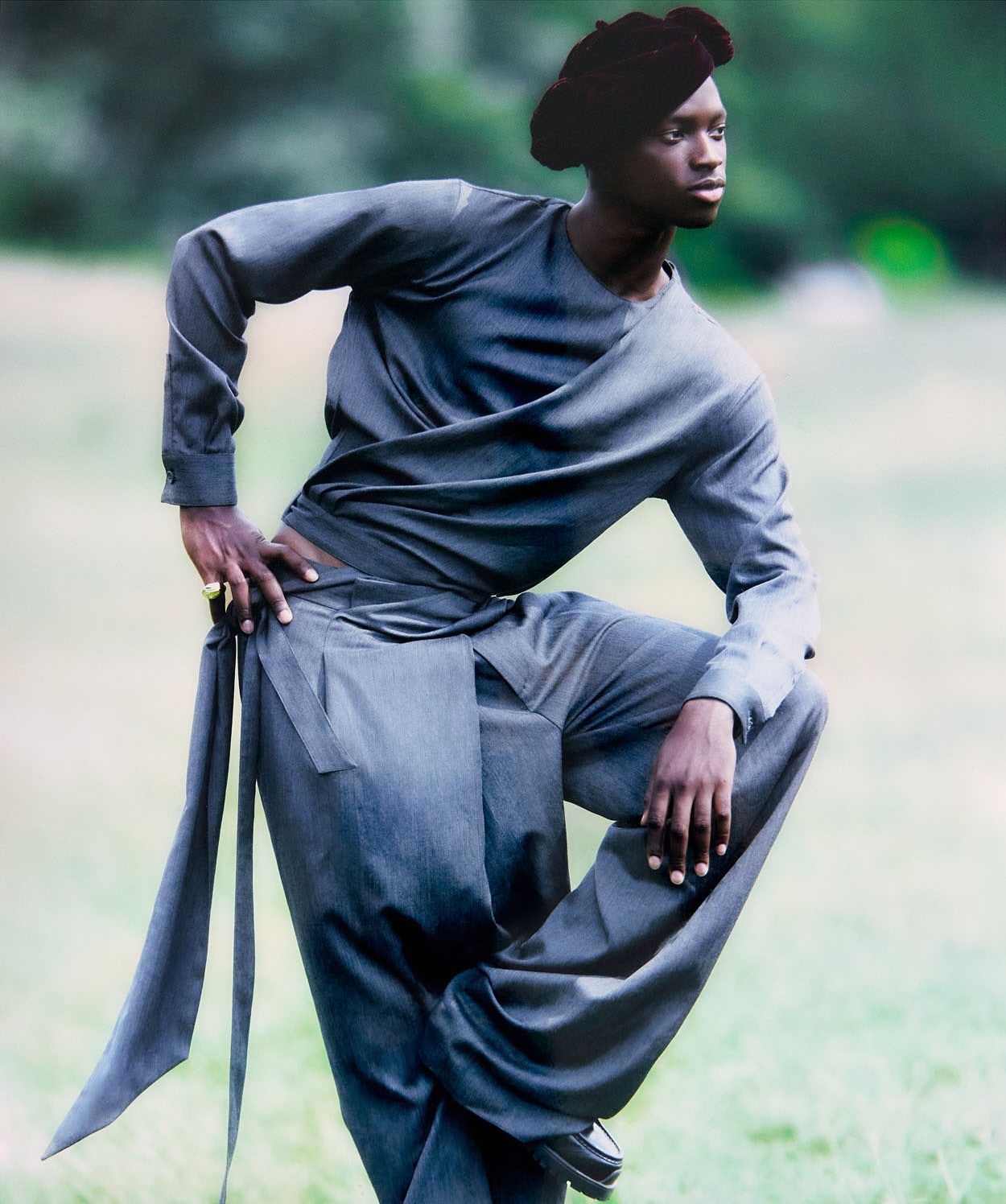 Fashionable person poses in a draped, long-sleeved grey outfit with a matching head covering in a grassy, outdoor setting.