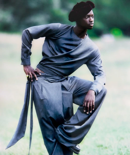 Fashionable person poses in a draped, long-sleeved grey outfit with a matching head covering in a grassy, outdoor setting.
