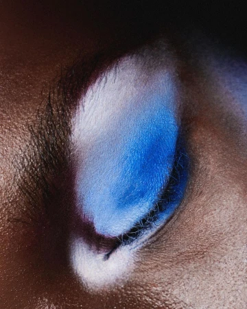 Close-up photo of an eye with blue and white eyeshadow on dark skin, with dark eyelashes and a blurry black background.
