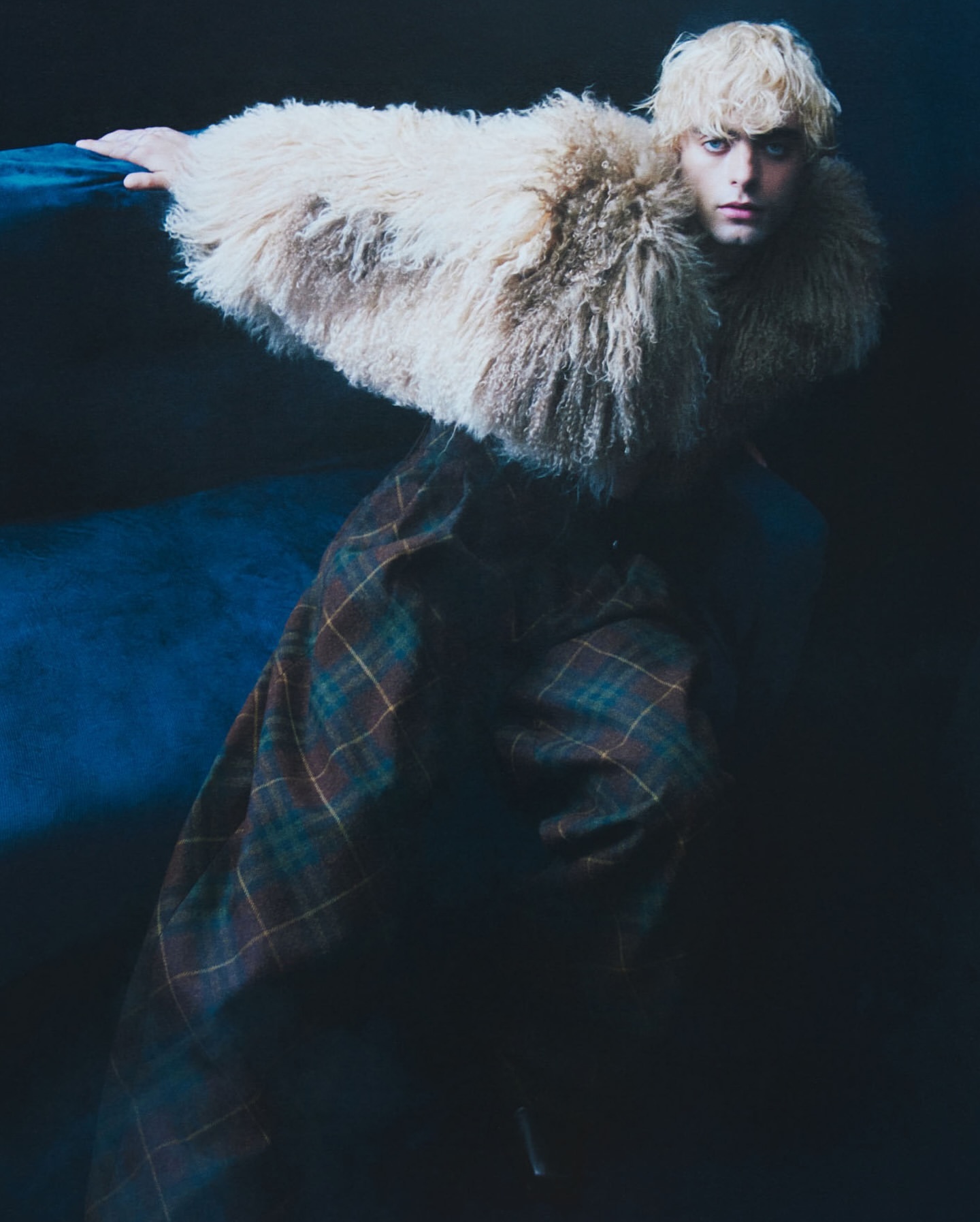 Fashion shot of a young adult with blonde hair wearing a large fur collar, plaid skirt, and posing in a dark studio with blue lighting.