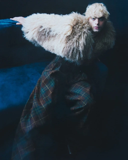 Fashion shot of a young adult with blonde hair wearing a large fur collar, plaid skirt, and posing in a dark studio with blue lighting.