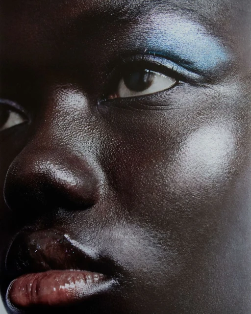 Close-up portrait of a dark-skinned woman with iridescent blue eyeshadow, glossy lips, and luminous highlighting. Beauty and makeup focus.
