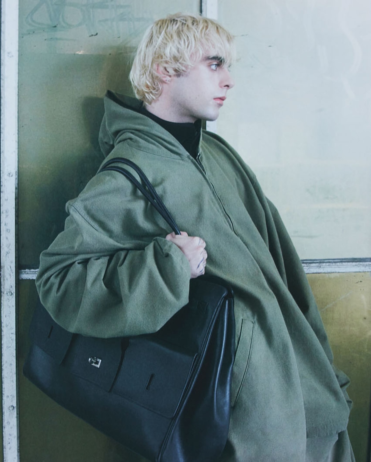 Blonde man wearing an oversized green hooded jacket and holding a large black leather shoulder bag in London