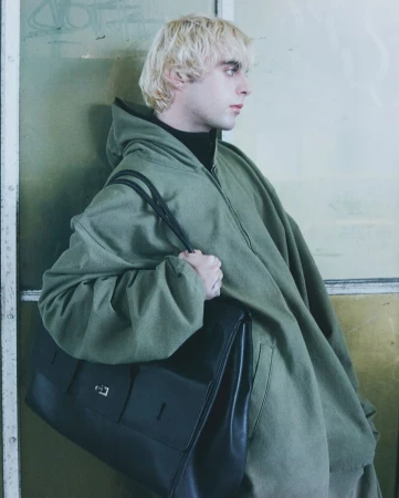 Blonde man wearing an oversized green hooded jacket and holding a large black leather shoulder bag in London