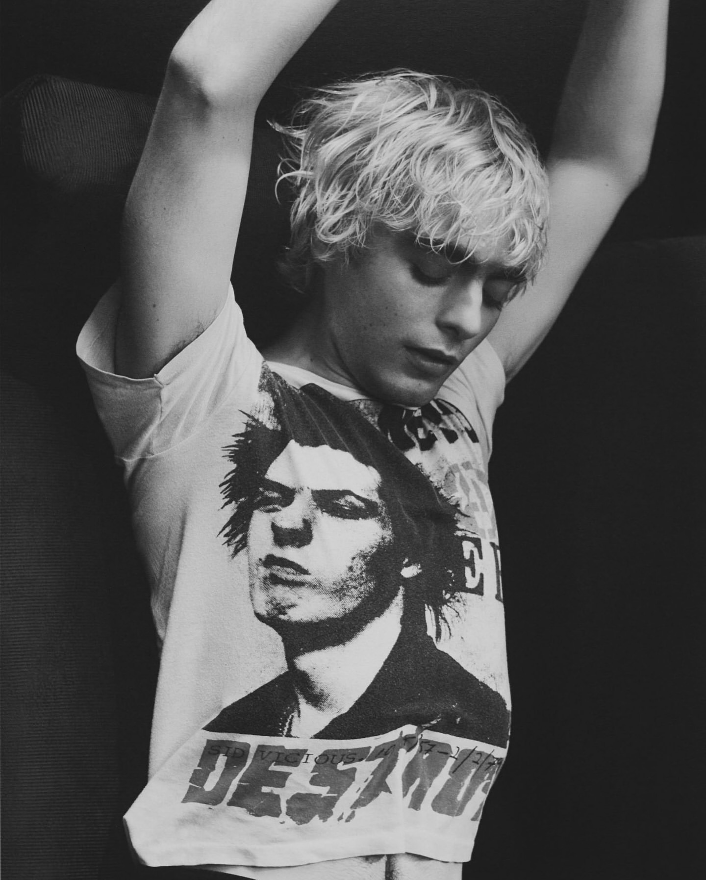 A young adult with blond hair poses indoors wearing a band tee shirt with arms above head in black and white, looking down.