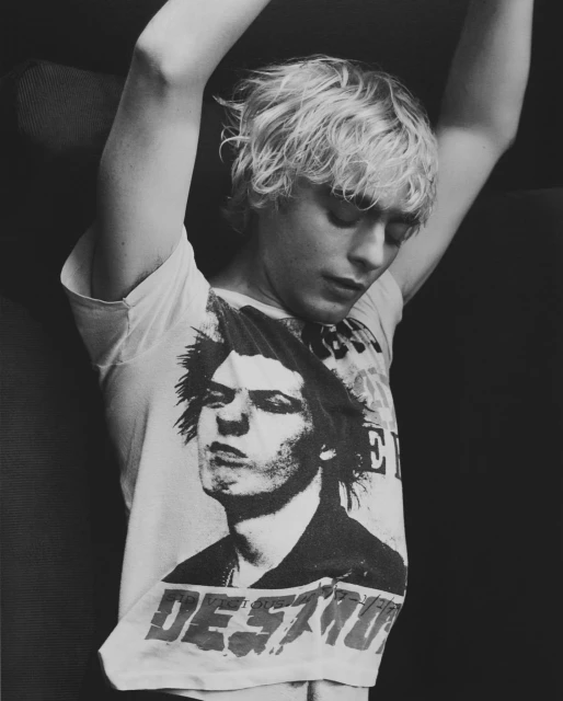 A young adult with blond hair poses indoors wearing a band tee shirt with arms above head in black and white, looking down.