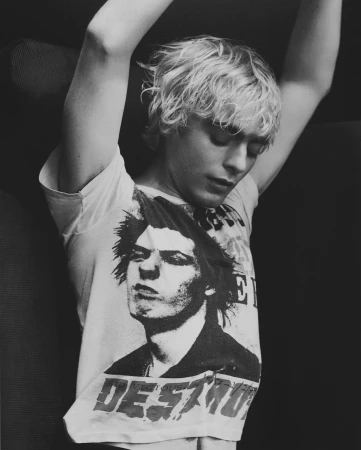 A young adult with blond hair poses indoors wearing a band tee shirt with arms above head in black and white, looking down.