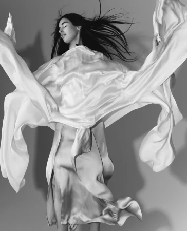 Model with long dark hair in a flowing white silk garment against a grey studio background captured in monochrome.