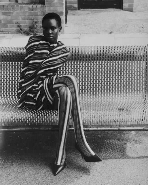 Black and white portrait of an adult female model with short hair sitting on a metal bench wearing striped top, tights, and high heels.