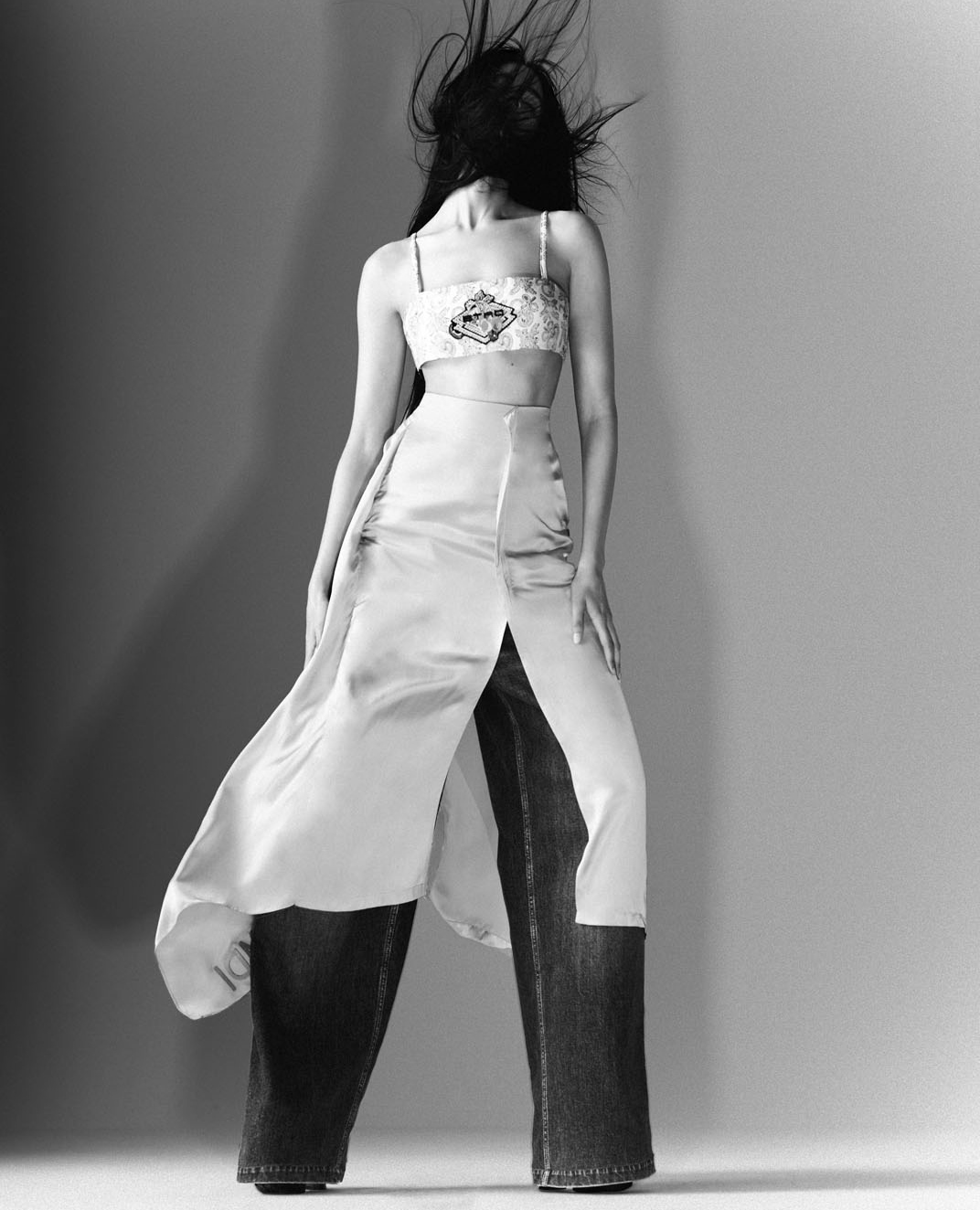 Monochrome fashion portrait: a slender woman with wind-swept hair poses in a cropped top, satin skirt, and wide-leg denim pants against a grey backdrop.