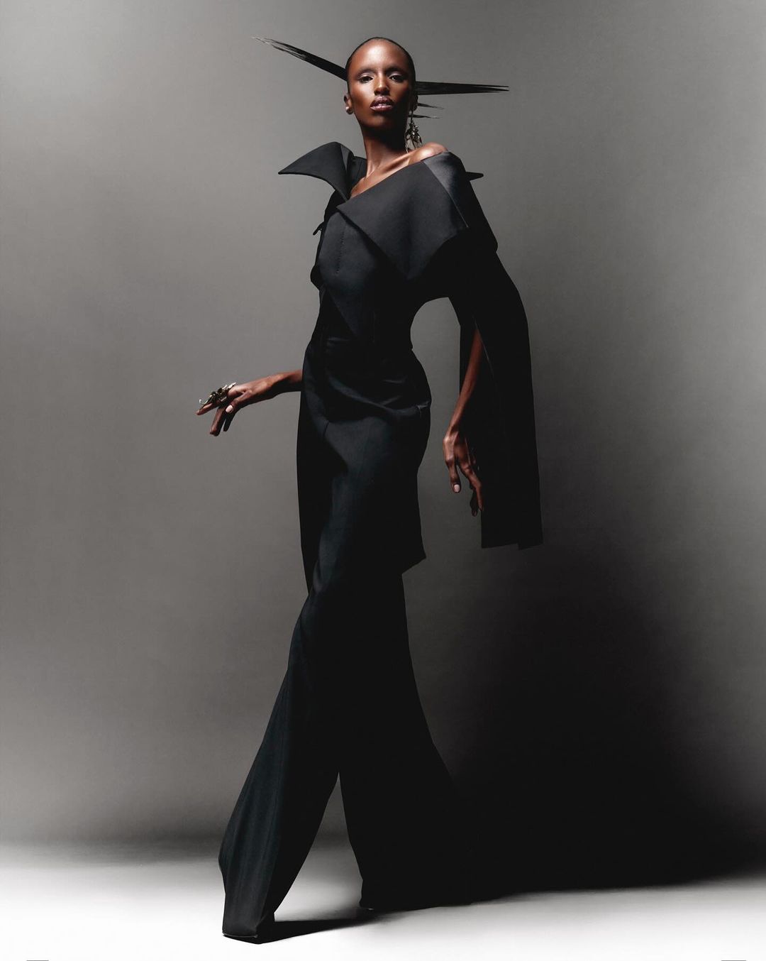 A fashion shot of a slender Black woman in a dramatic black outfit posing against a grey backdrop, showcasing avant-garde style.