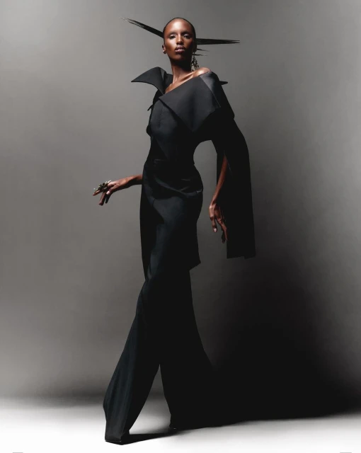 A fashion shot of a slender Black woman in a dramatic black outfit posing against a grey backdrop, showcasing avant-garde style.
