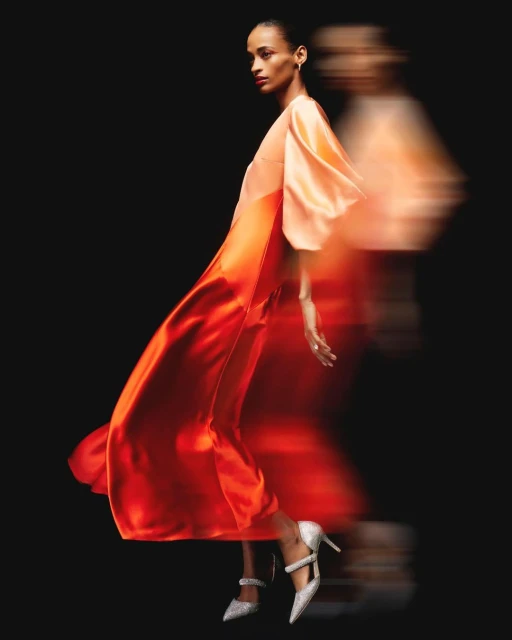 Elegant fashion shot of an adult woman in motion wearing a long orange dress and high heels against a dark black background in studio.