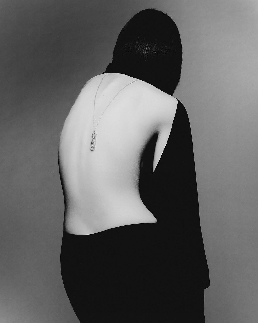 Back view of a woman with bob haircut wearing a black top and necklace, captured in a monochrome studio shot.