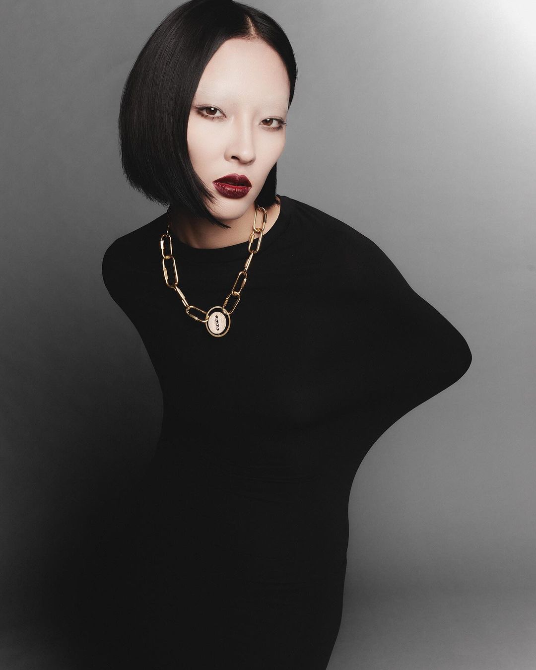 A person with pale skin and a dark bob haircut poses in a studio wearing a black dress and gold necklace against a grey backdrop.