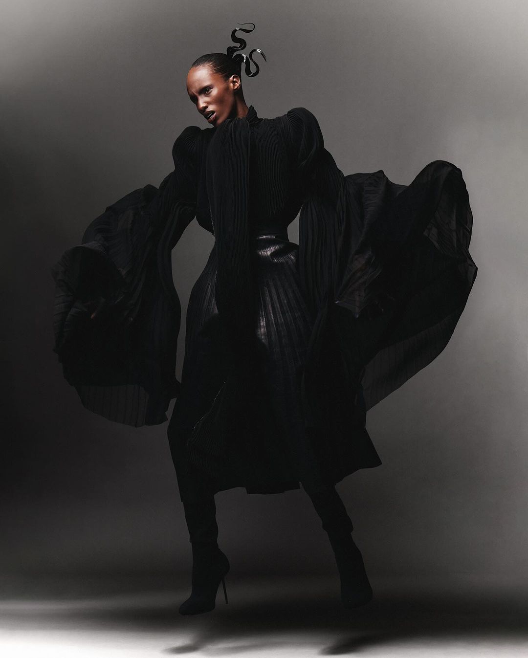 A striking full-length fashion shot of a Black female model with dark hair in an elaborate black gown and heels against a grey backdrop.