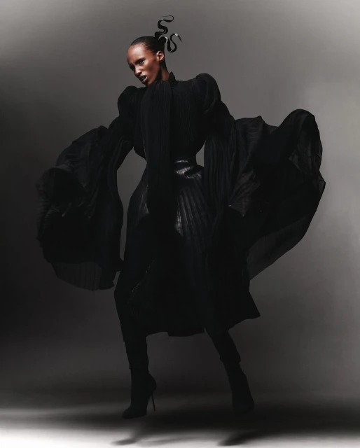 A striking full-length fashion shot of a Black female model with dark hair in an elaborate black gown and heels against a grey backdrop.