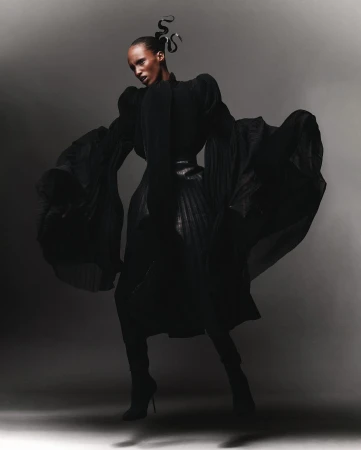 A striking full-length fashion shot of a Black female model with dark hair in an elaborate black gown and heels against a grey backdrop.