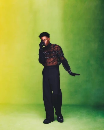 A man in a black sheer sequined top and dark trousers posing against a yellow and green gradient backdrop.