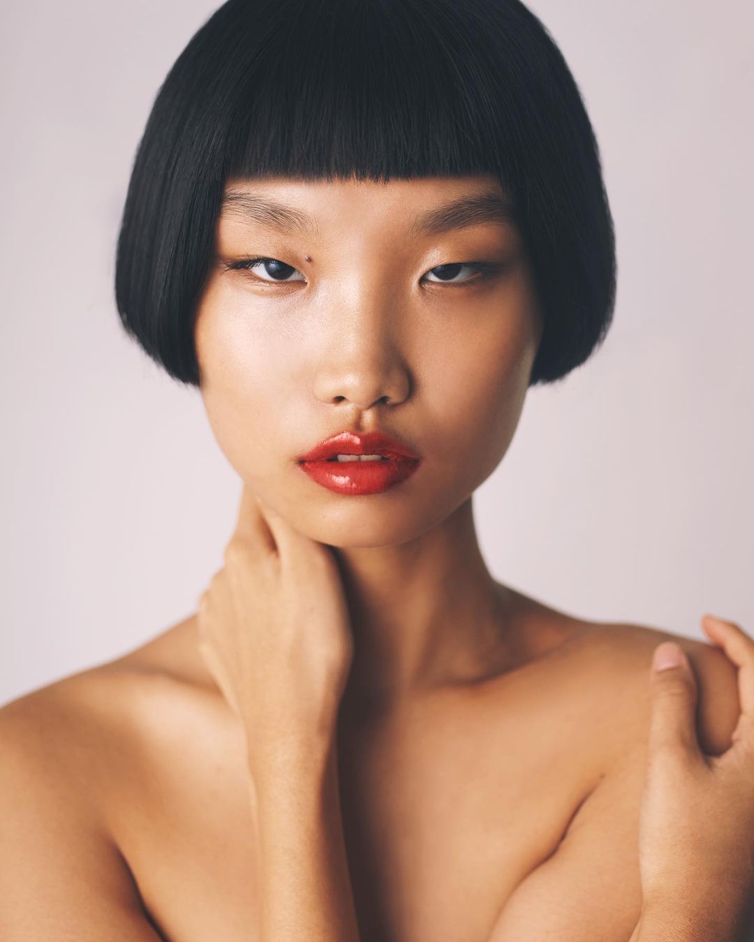 Close-up portrait of a model with a blunt bob haircut and red lipstick, against a neutral background, emphasizing beauty and fashion.
