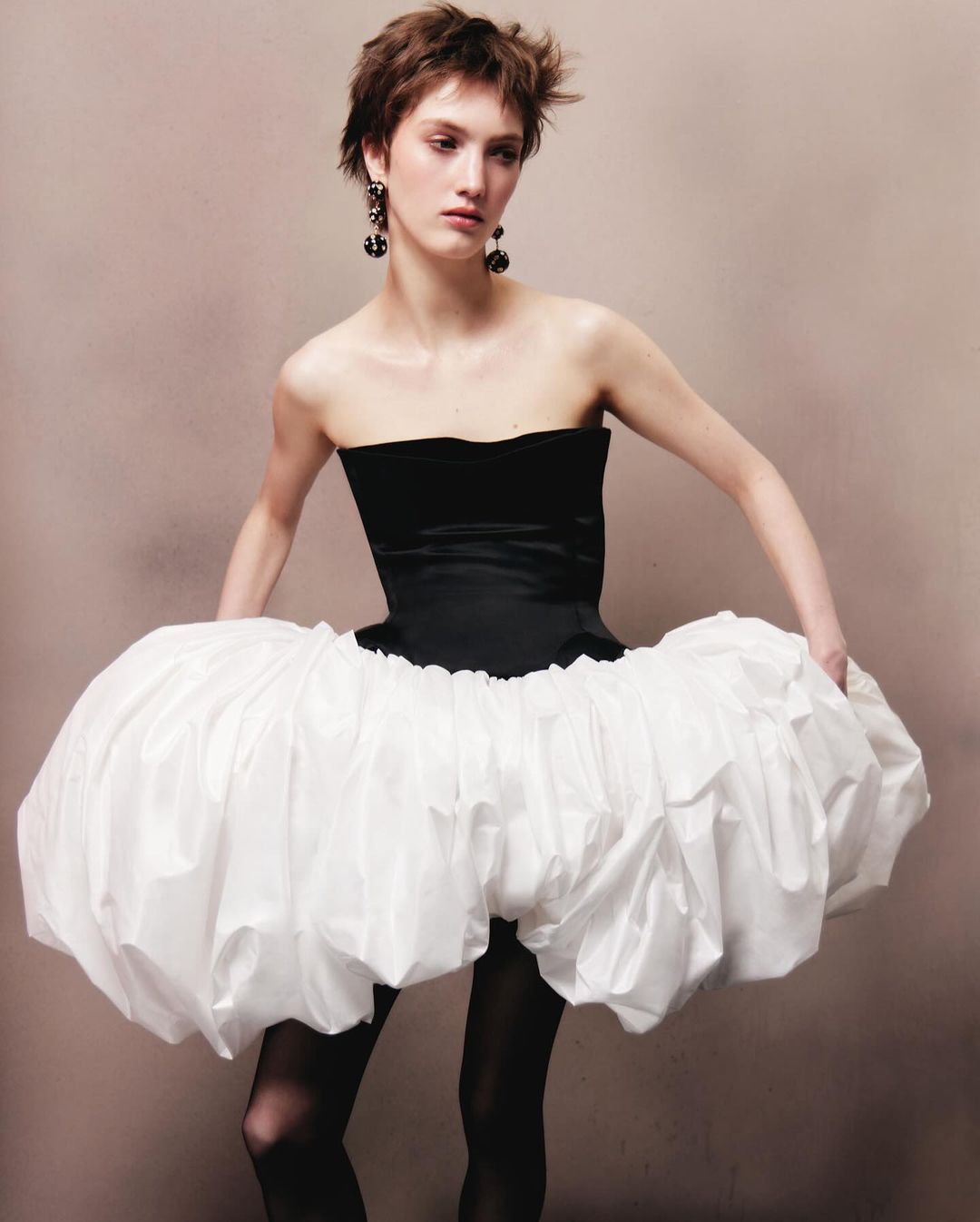 Model with short brown hair wearing a black strapless bodice and voluminous white puff skirt against a plain grey background