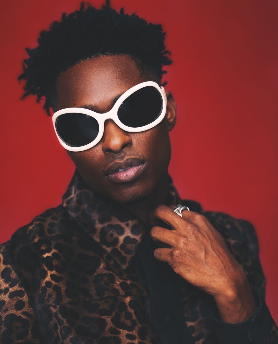 Stylish portrait of a Black man with sunglasses and leopard-print jacket, set against a red background. Fashion and celebrity style.