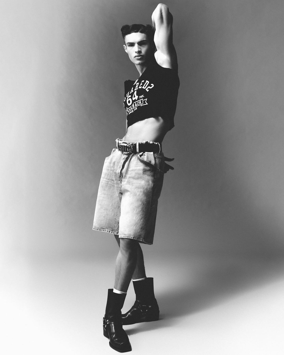 Monochrome portrait of a male model wearing a cropped graphic t-shirt and denim shorts against a grey seamless backdrop.