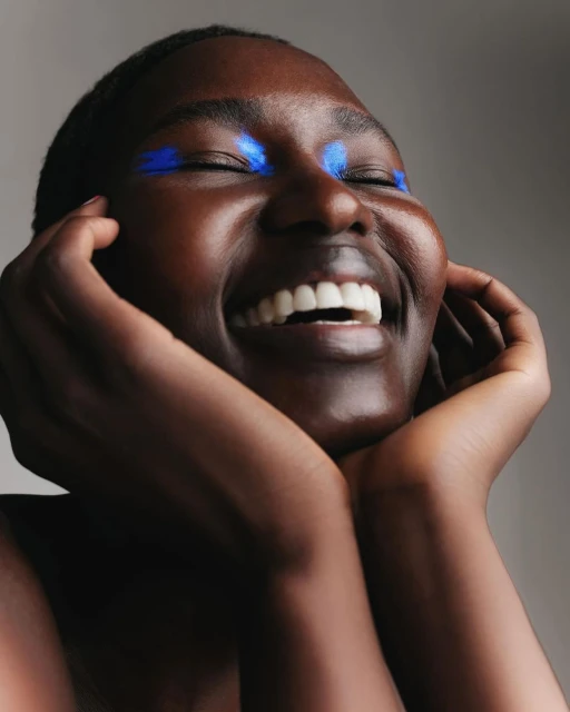 Close-up of a smiling African woman with closed eyes and blue makeup around her eyes, placing her hands on her cheeks in an indoor setting.