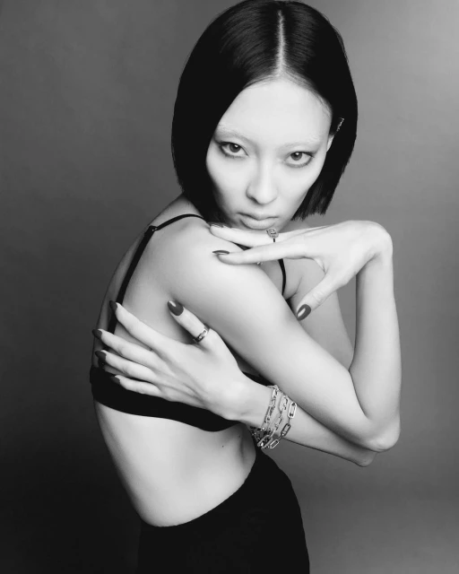 Black-and-white portrait of an adult female with light skin, short black hair, and a dark bra, posing with hands near the shoulder.